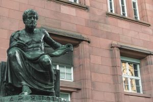 Estatua de Aristóteles sentado con un pergamino en la mano frente a un edificio de piedra roja