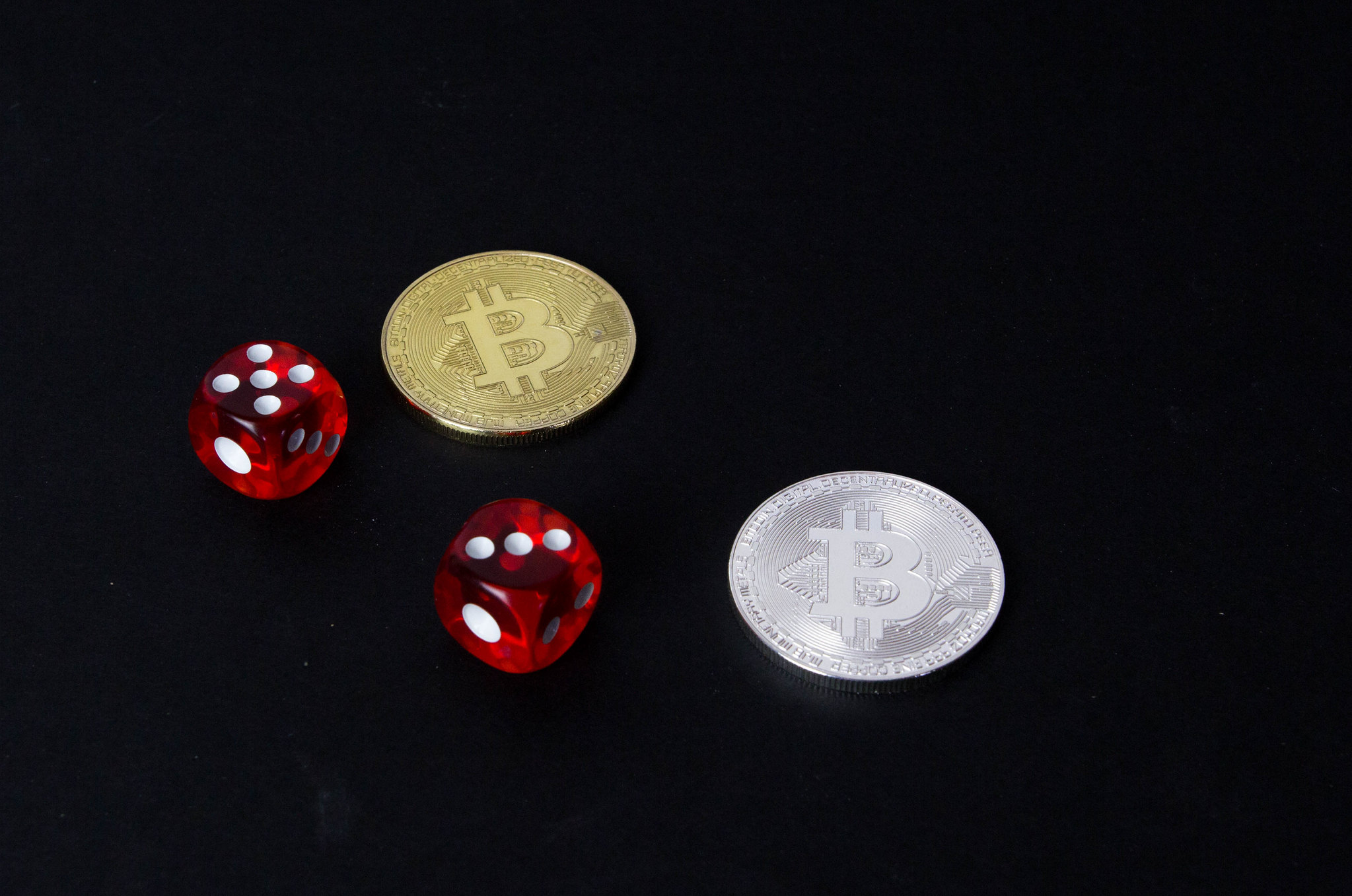 Monedas de Bitcoin doradas y plateadas junto a dos dados rojos sobre fondo negro.