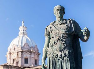 Estatua de Julio César en bronce con toga y armadura romana, situada frente a la cúpula de la iglesia de los Santos Luca y Martina en Roma, representando el poder político y militar de la antigua Roma