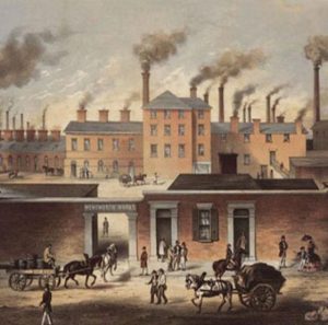Ilustración de una fábrica del siglo XIX durante la Revolución Industrial, con numerosos edificios de ladrillo, chimeneas expulsando humo, carruajes tirados por caballos y trabajadores entrando y saliendo del recinto