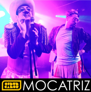 jete Calor en concierto con atuendos extravagantes: uno de los integrantes canta con sombrero blanco, gafas y chaqueta con hombreras doradas; el otro posa con actitud mientras lleva una chaqueta de flecos y minifalda. Estética kitsch y humor absurdo que define el estilo del grupo y su sátira del concepto 'mocatriz