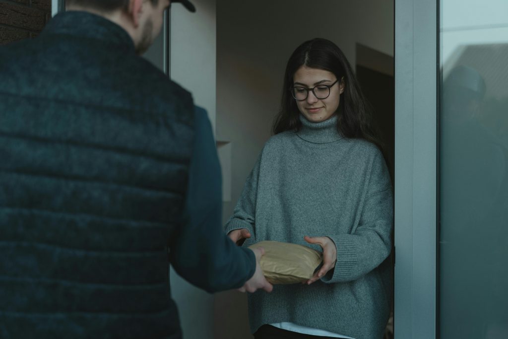 Mujer joven con gafas y suéter gris recibe un paquete de un repartidor en la puerta de su casa