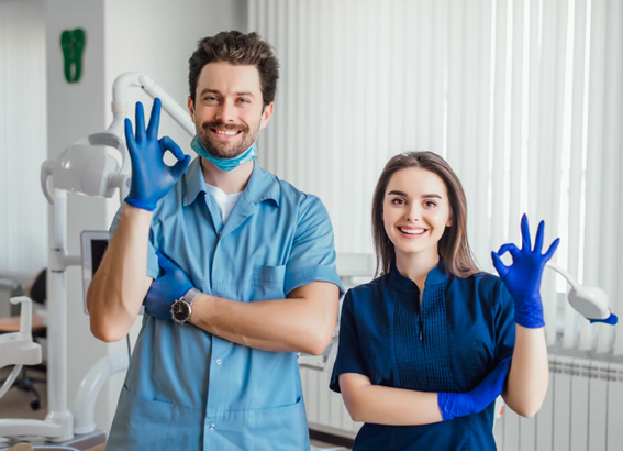 Dos profesionales de la salud sonríen en una clínica dental, llevando guantes azules y haciendo el gesto de ‘ok’ con la mano.