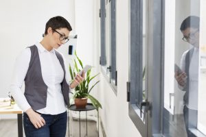 Mujer en un entorno de oficina leyendo su teléfono móvil junto a una ventana, vestida con camisa blanca, chaleco gris y gafas.