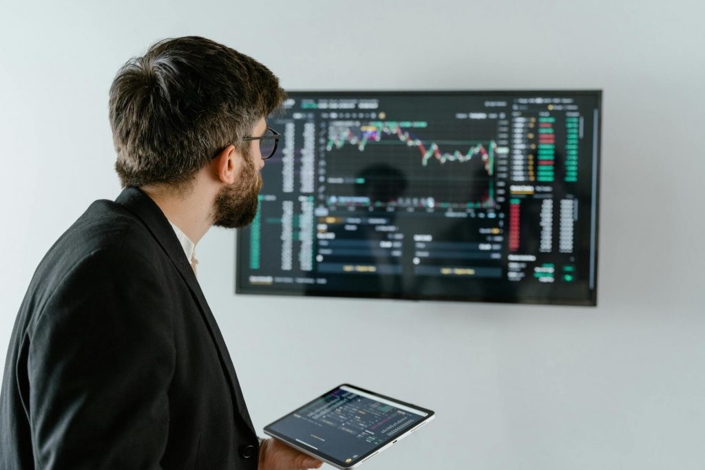 Hombre analizando gráficos bursátiles en una pantalla mientras sostiene una tablet con datos de trading financiero.
