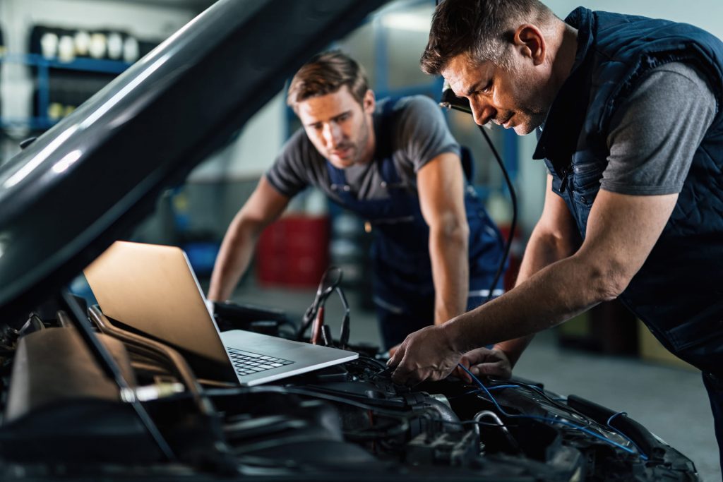 Dos mecánicos trabajando en el motor de un coche en un taller, utilizando un ordenador portátil para el diagnóstico del vehículo.