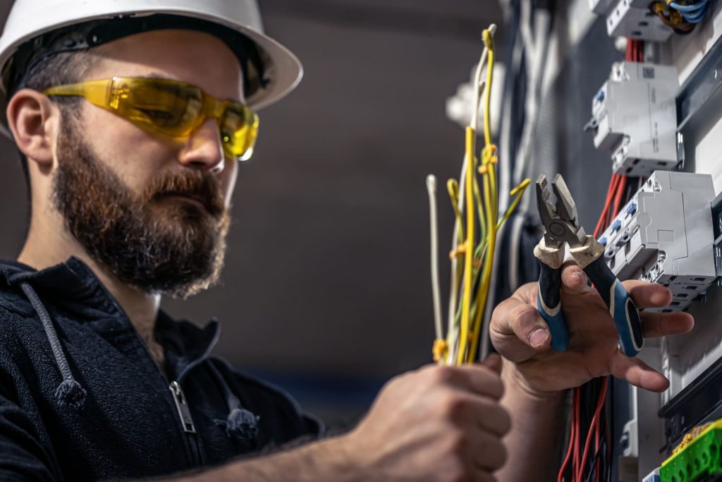Electricista con casco y gafas de seguridad trabajando en un cuadro eléctrico, sujetando cables y usando alicates para realizar una instalación o mantenimiento.