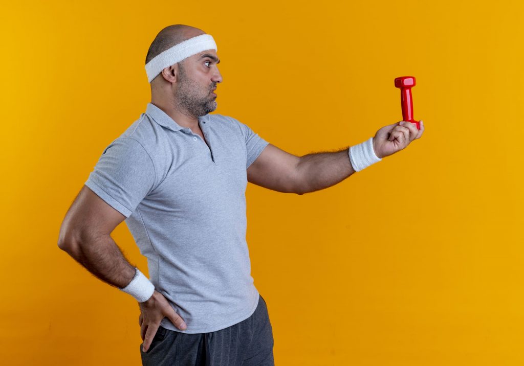 Hombre con ropa deportiva sosteniendo una mancuerna pequeña con gesto de duda frente a un fondo amarillo.