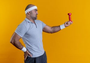 Hombre con ropa deportiva sosteniendo una mancuerna pequeña con gesto de duda frente a un fondo amarillo.