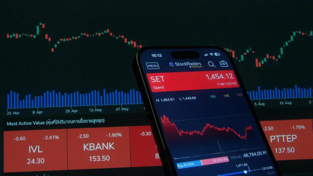 Teléfono móvil mostrando una aplicación de bolsa con gráfico en descenso frente a un monitor con cotizaciones y velas bursátiles en rojo.