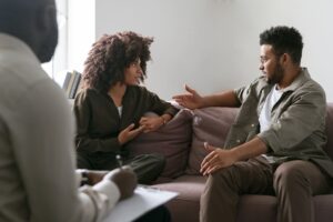 Pareja sentada en un sofá durante una sesión de terapia, conversando seriamente mientras un terapeuta toma notas en primer plano.