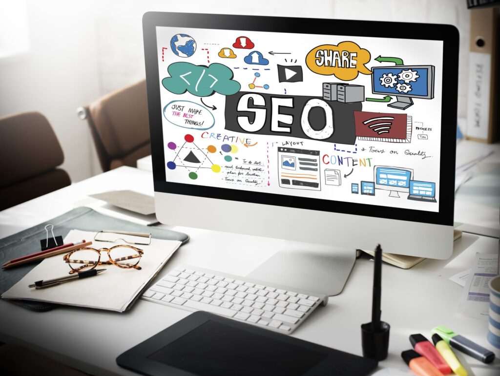 Monitor de escritorio mostrando la palabra SEO con iconos de contenido, analítica, nube y diseño web en un entorno de trabajo con teclado, tableta gráfica y material de oficina
