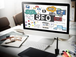 Monitor de escritorio mostrando la palabra SEO con iconos de contenido, analítica, nube y diseño web en un entorno de trabajo con teclado, tableta gráfica y material de oficina