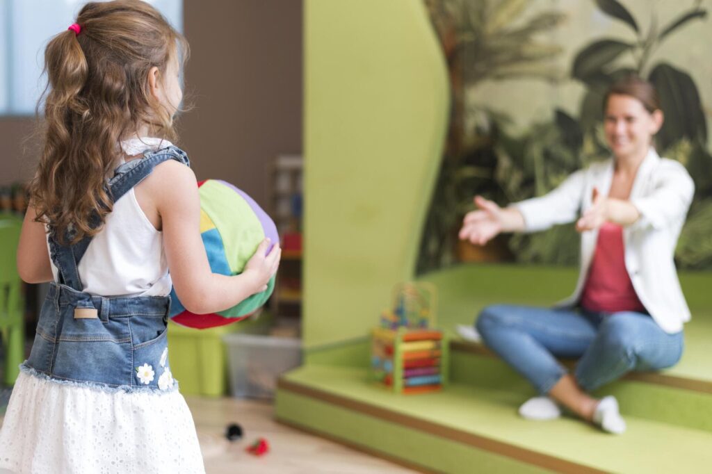 Niña pequeña sosteniendo una pelota de colores frente a una terapeuta sentada con los brazos abiertos en una sala de juegos infantil