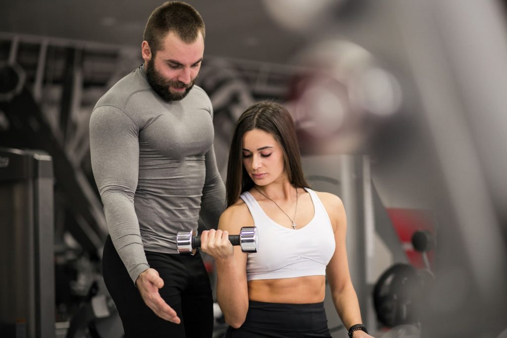 Entrenador personal supervisando a una mujer mientras realiza un ejercicio de bíceps con mancuernas en el gimnasio.