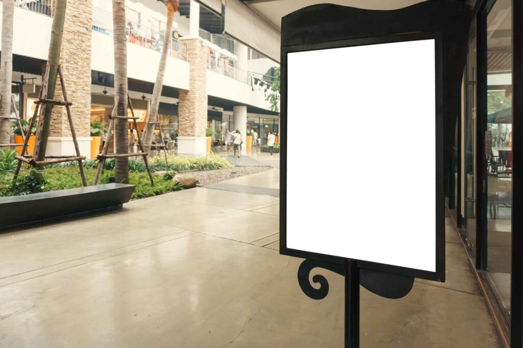 ótem publicitario con pantalla en blanco en un centro comercial abierto, con palmeras, tiendas y personas caminando al fondo.