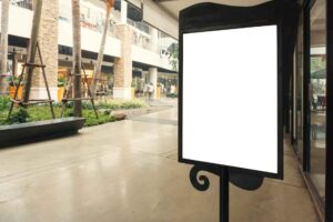 ótem publicitario con pantalla en blanco en un centro comercial abierto, con palmeras, tiendas y personas caminando al fondo.