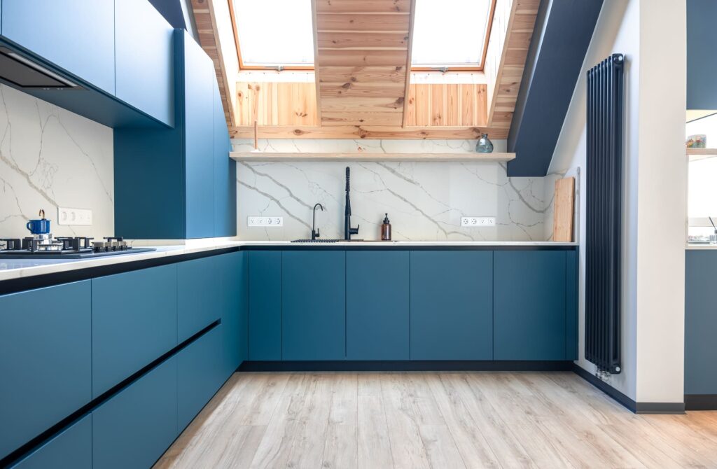 Cocina moderna en forma de L con muebles azules, encimera blanca efecto mármol, grifo negro y techo abuhardillado con vigas de madera y ventanas tipo Velux.