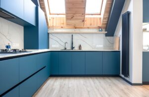 Cocina moderna en forma de L con muebles azules, encimera blanca efecto mármol, grifo negro y techo abuhardillado con vigas de madera y ventanas tipo Velux.