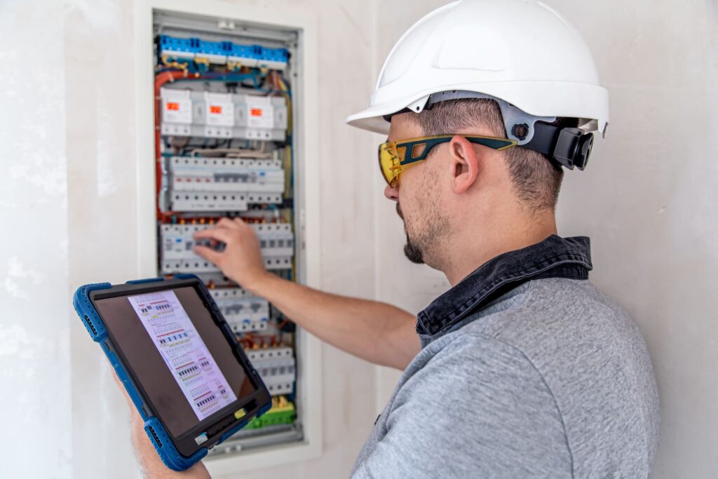 Electricista con casco y gafas de seguridad revisando un cuadro eléctrico mientras consulta el esquema en una tablet.