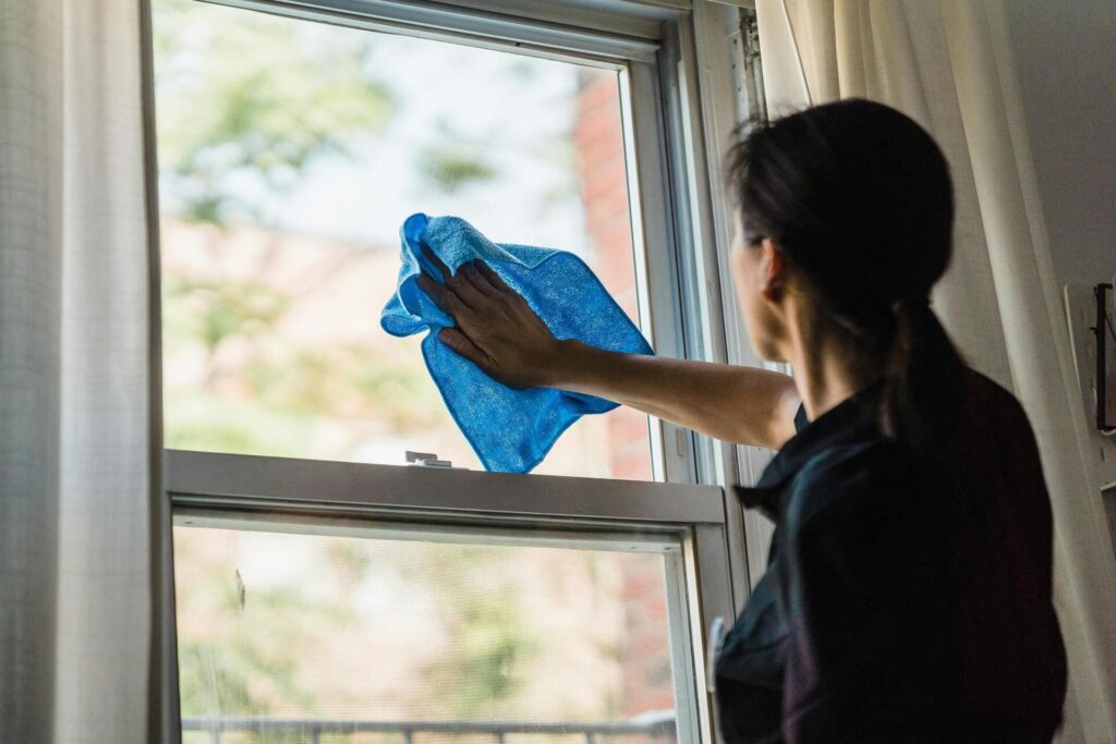 Mujer limpiando el cristal de una ventana con un paño azul en el interior de una vivienda.