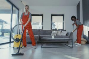 Dos trabajadoras de limpieza con uniforme rojo limpiando un salón moderno, una aspirando el suelo y otra fregando cerca del sofá.