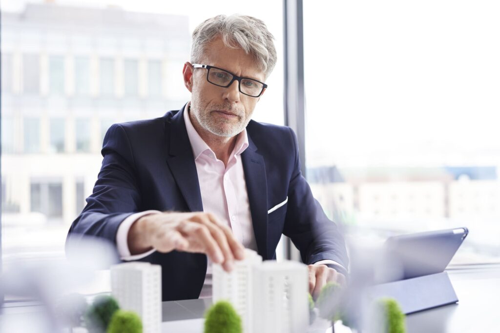 Hombre de negocios con gafas revisando una maqueta de edificios sobre una mesa en una oficina luminosa, con una tablet al lado.
