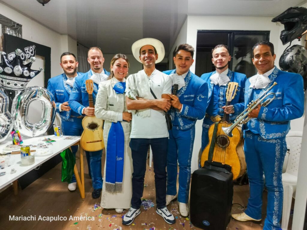 Grupo de mariachis vestidos con trajes azules y una mujer con traje tradicional, posando junto a un hombre con sombrero en una celebración interior con instrumentos musicales y decoración festiva.