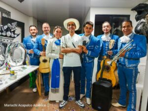 Grupo de mariachis vestidos con trajes azules y una mujer con traje tradicional, posando junto a un hombre con sombrero en una celebración interior con instrumentos musicales y decoración festiva.