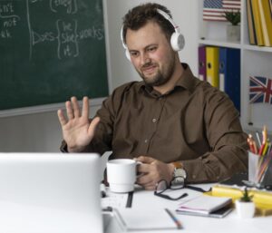 Profesor dando una clase de inglés online con auriculares y saludando a través del ordenador mientras está sentado en su escritorio frente a una pizarra.