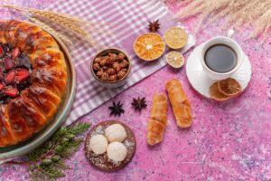 Desayuno con café en taza blanca, rosca dulce rellena de fresas y chocolate, galletas enrolladas, coco rallado y frutos secos sobre mesa rosa decorada con cítricos y especias.