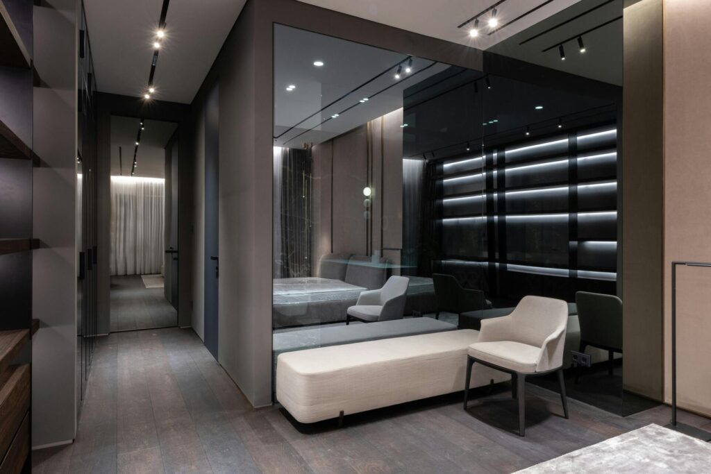 Dormitorio moderno con pared de cristal, iluminación indirecta y zona de descanso con banco y sillón junto a la cama.