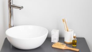 Lavabo moderno con grifo cromado sobre encimera, acompañado de cepillos de dientes de bambú, cepillo de madera, bloque de mármol y frasco de aceite en un entorno minimalista.