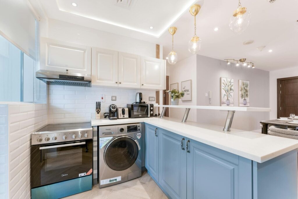 Cocina moderna de estilo luminoso con muebles blancos y azules, encimera en forma de L, horno, campana extractora y lavadora integrados, con iluminación colgante decorativa.