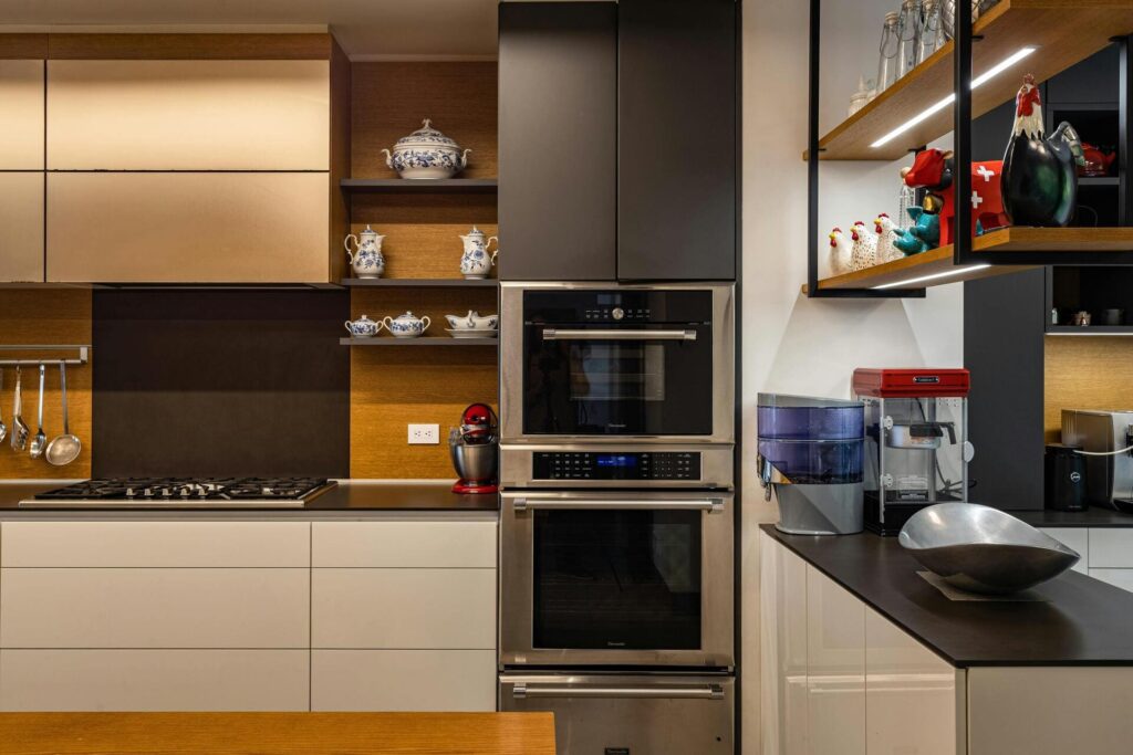 Cocina moderna con mobiliario en tonos madera y blanco, equipada con horno empotrado doble, encimera de gas, estanterías decorativas y pequeños electrodomésticos sobre la encimera.