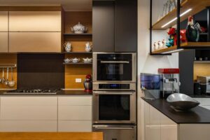 Cocina moderna con mobiliario en tonos madera y blanco, equipada con horno empotrado doble, encimera de gas, estanterías decorativas y pequeños electrodomésticos sobre la encimera.