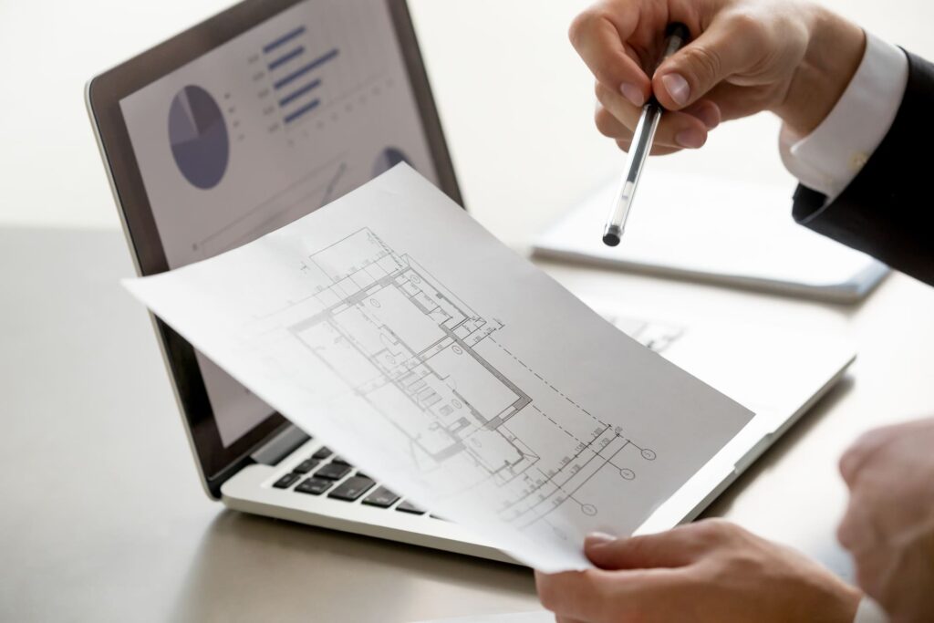 Mano sosteniendo un plano arquitectónico mientras señala detalles con un bolígrafo frente a un portátil con gráficos de análisis.