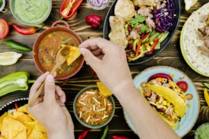 Mesa con platos de comida mexicana, tacos, nachos, salsas y verduras, mientras una persona sirve salsa con una cuchara sobre un totopo.