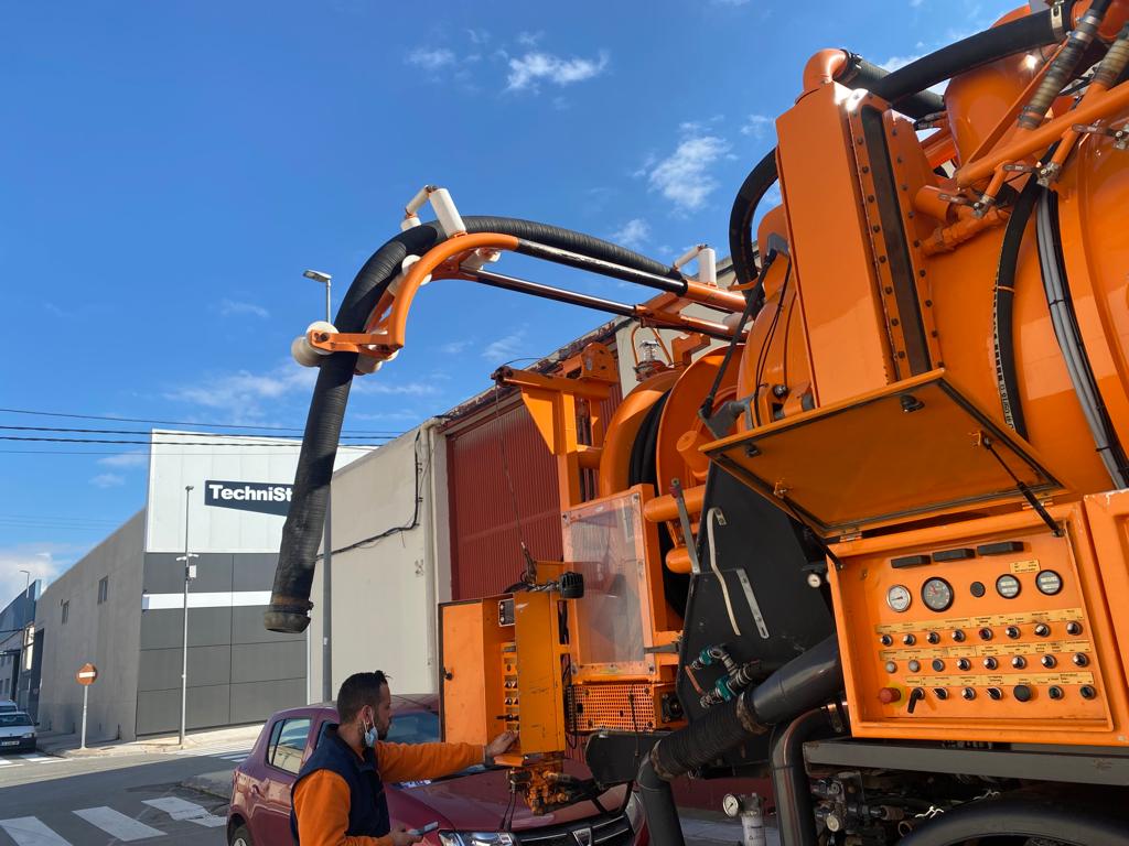 Camión cuba naranja para desatascos con manguera de succión desplegada junto a un operario en una zona industrial.