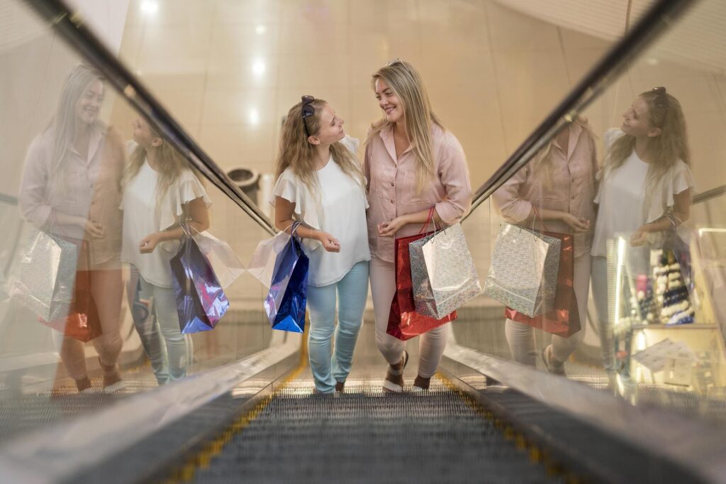 Dos mujeres sonrientes con bolsas de compras subiendo por unas escaleras mecánicas en un centro comercial.