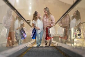 Dos mujeres sonrientes con bolsas de compras subiendo por unas escaleras mecánicas en un centro comercial.