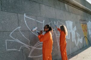 Dos personas con monos naranjas pintando sobre un muro de piedra para cubrir grafitis.