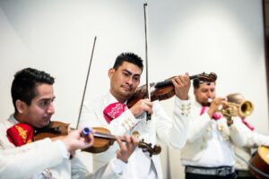 Grupo de mariachis con trajes blancos tocando violines y trompeta durante una actuación musical.