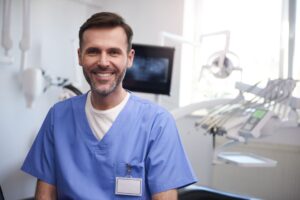 Dentista sonriente con uniforme sanitario azul en una clínica dental moderna equipada con sillón odontológico e instrumental profesional al fondo.