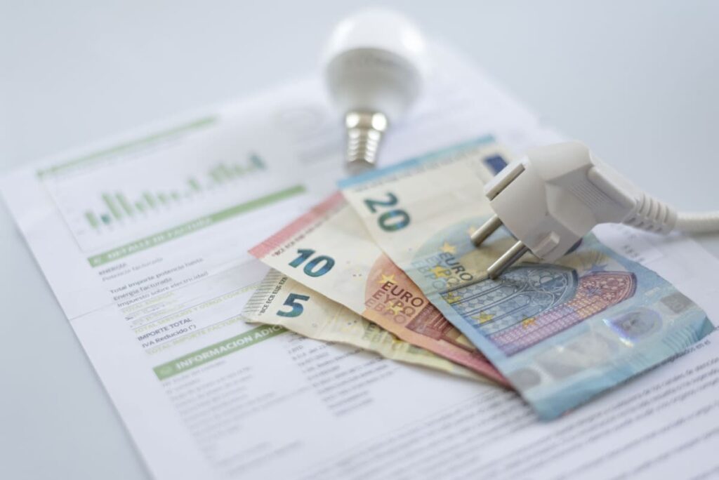 Factura de electricidad con un enchufe, una bombilla LED y varios billetes de euro sobre ella, representando el coste del consumo eléctrico y el pago de la luz.