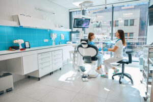 Dentista en Segur de Calafell atendiendo a una paciente en el sillón dental de la consulta, con pared de azulejos azules, monitor de pantalla y ventanales con vistas a la ciudad.