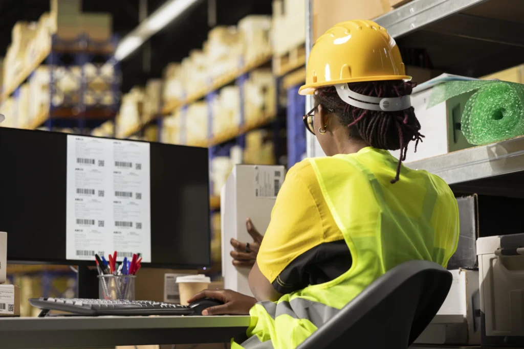 Trabajadora de logística con casco y chaleco reflectante imprimiendo etiquetas de seguimiento para paquetes en un almacén.