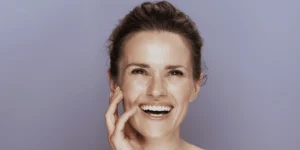 Mujer sonriente tocándose el rostro sobre un fondo neutro, mostrando una piel cuidada y luminosa.