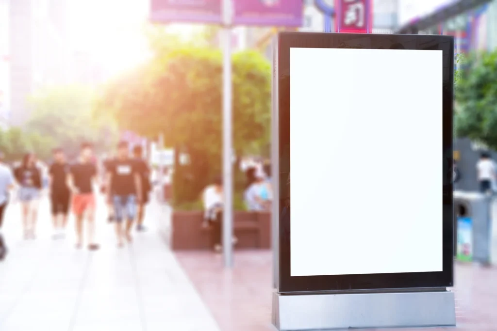 Tótem publicitario con pantalla en blanco situado en una calle concurrida con peatones desenfocados al fondo